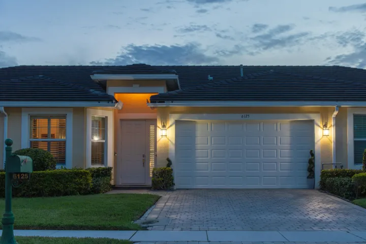 Exterior featuring a paver driveway, an attached garage, a covered entry, exterior lighting, and manicured shrubbery
