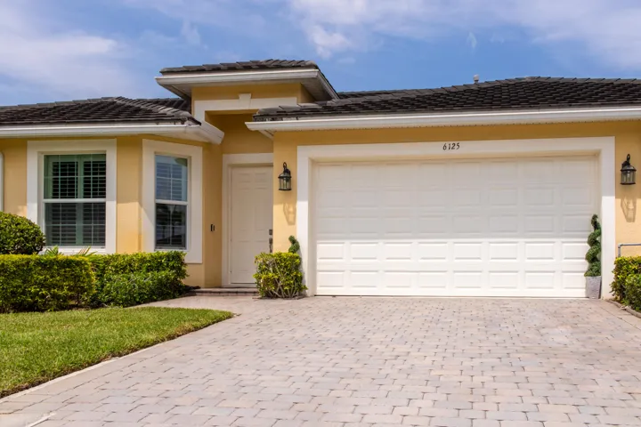 Cream stucco exterior with a dark tile roof, featuring a paver driveway, an attached garage, a panel-style front door, and decorative outdoor lighting fixtures