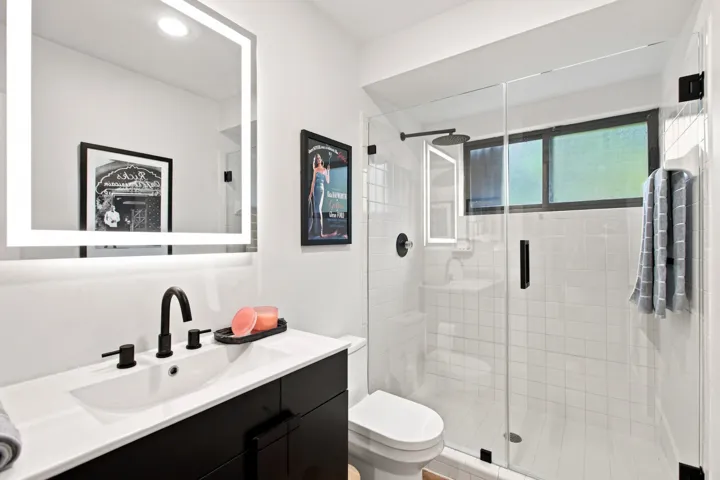 Modern bathroom featuring a rectangular vanity with a white countertop and black cabinetry, a backlit mirror, a walk-in shower with clear glass enclosure, and a black rain shower head