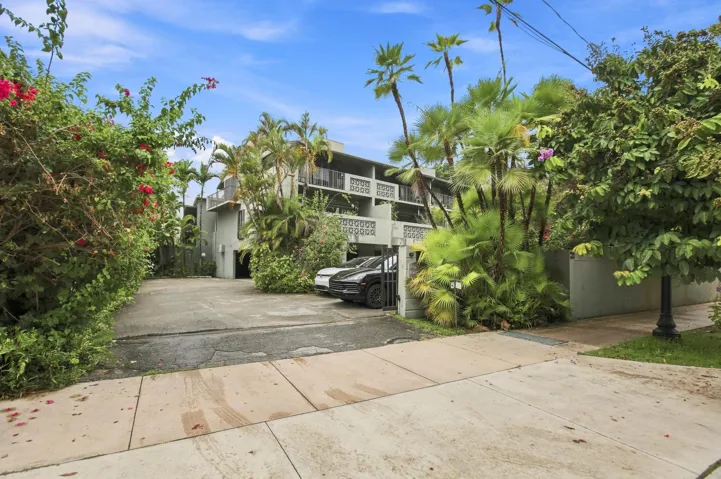 Multi-story building featuring exterior balconies with decorative screens, lush tropical landscaping, and a paved driveway leading to covered parking