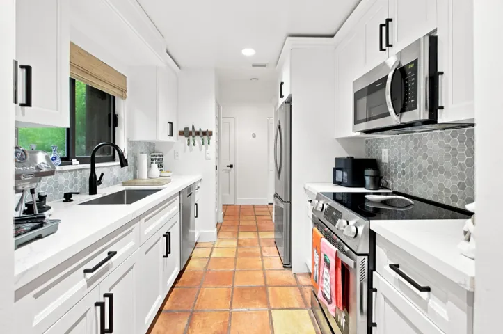 Galley kitchen featuring white cabinetry, stainless steel appliances, a hexagon tile backsplash, light-colored countertops, and terracotta tile flooring