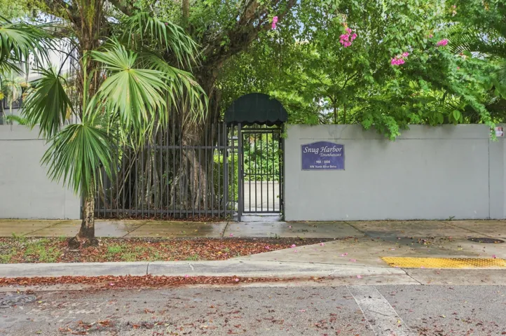Decorative iron gate entrance with a dark green awning, flanked by a concrete perimeter wall and mature tropical foliage