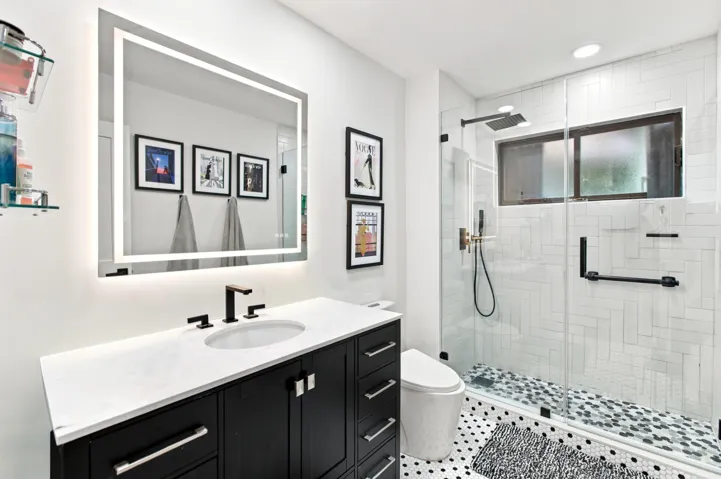 Bathroom featuring a black vanity with a white countertop, an illuminated mirror, a glass-enclosed shower with white subway tile, and a pebble-style shower floor