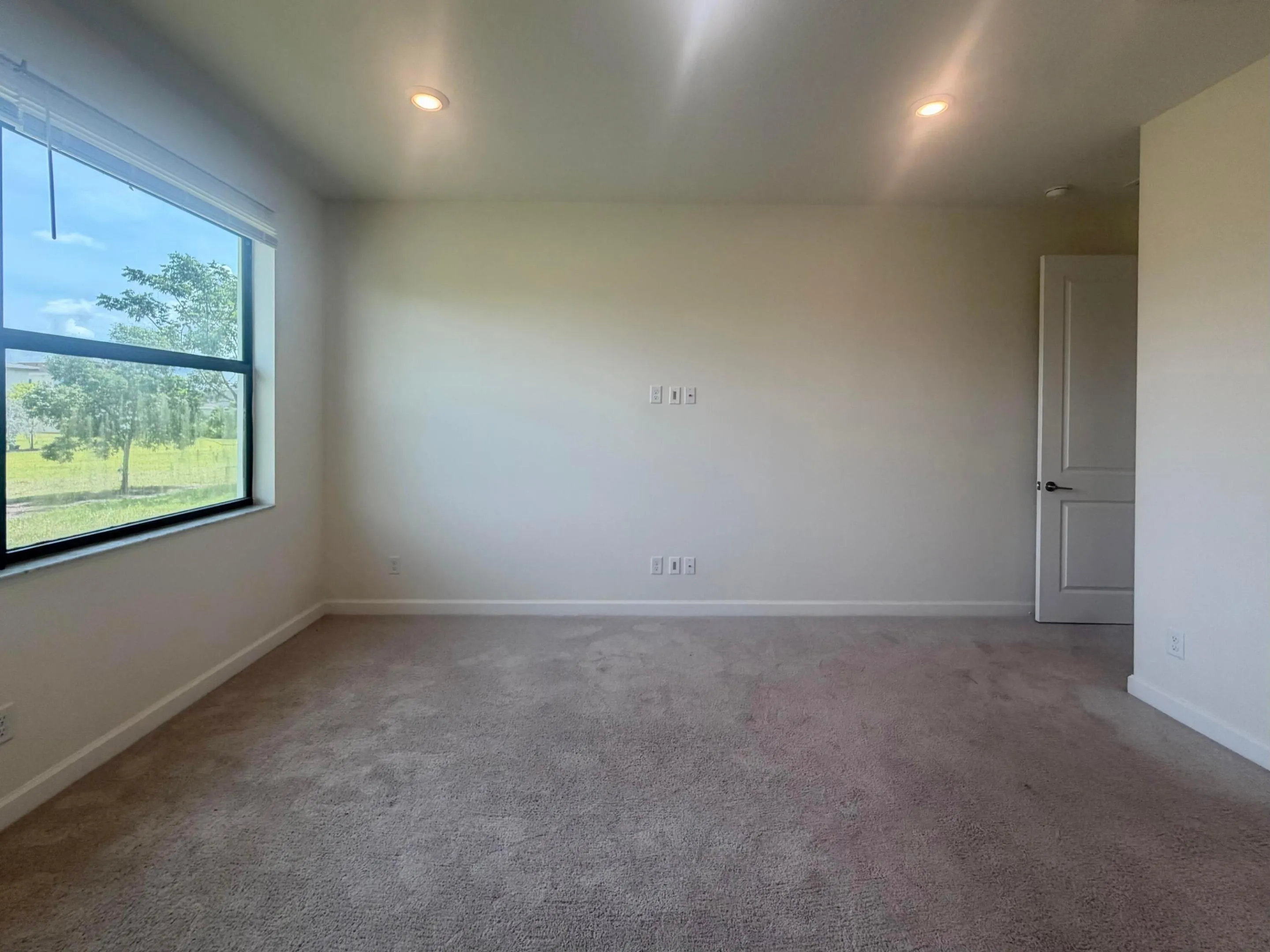 Bright interior space featuring a large window, neutral carpeting, recessed lighting, a paneled door, and white baseboards