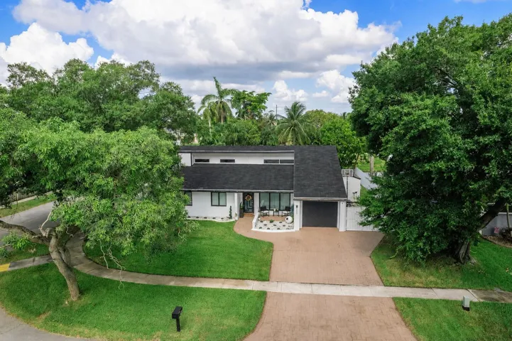 Modern white exterior with a dark shingle roof, a two-car garage, mature trees, and a paver driveway