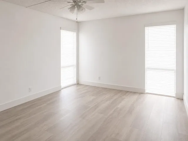 Spacious room with light wood-finish flooring, two large windows with horizontal blinds, and a ceiling fan
