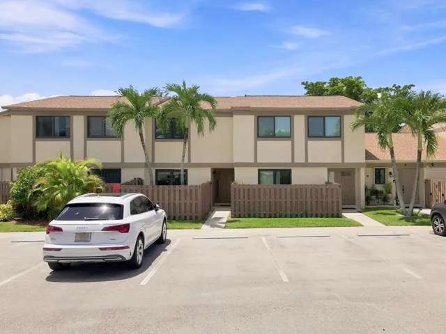 Two-story stucco exterior, brown shingle roof, brown privacy fencing, mature palm trees, and established landscaping