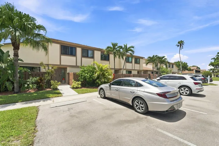 Two-story stucco exterior with contrasting trim, private fenced entrance, lush perimeter landscaping, and adjacent paved parking