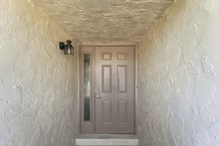 Textured stucco exterior with a paneled entry door, sidelight, and wall-mounted exterior light fixture