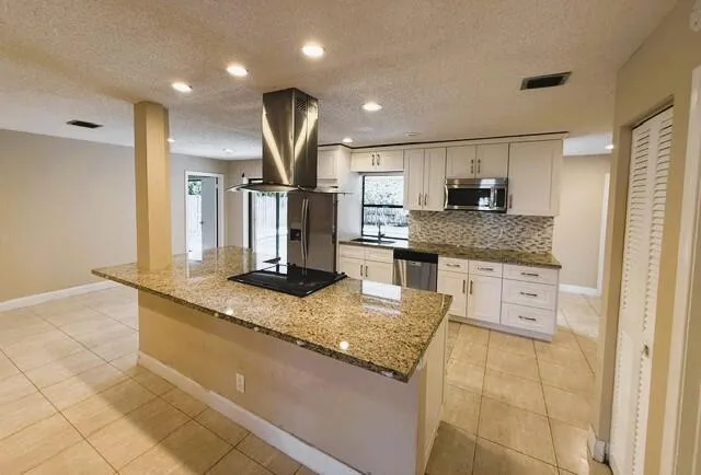 Kitchen featuring an island with a built-in cooktop and stainless steel vent hood, granite countertops, white cabinetry with silver hardware, stainless steel appliances, and tiled flooring