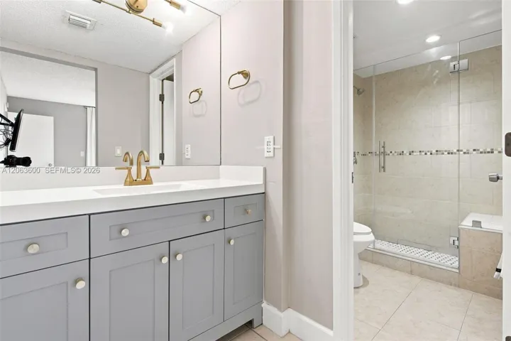 Ensuite bathroom with gray shaker vanity, quartz countertop, brass fixtures, and glass-enclosed shower beyond.