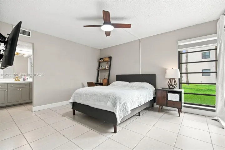 Primary bedroom with tile flooring, ceiling fan, wall-mounted TV, and direct access to the ensuite bathroom.