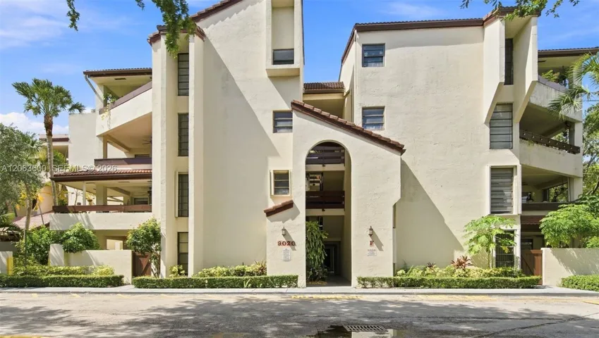 Front entrance to unit F, Spanish-tile roofline, arched breezeway, and mature landscaping with tropical foliage.