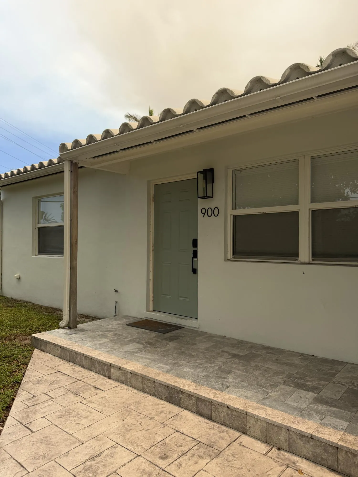 Front entry featuring a light green door, black exterior sconce, and stamped concrete patio leading to a tiled porch