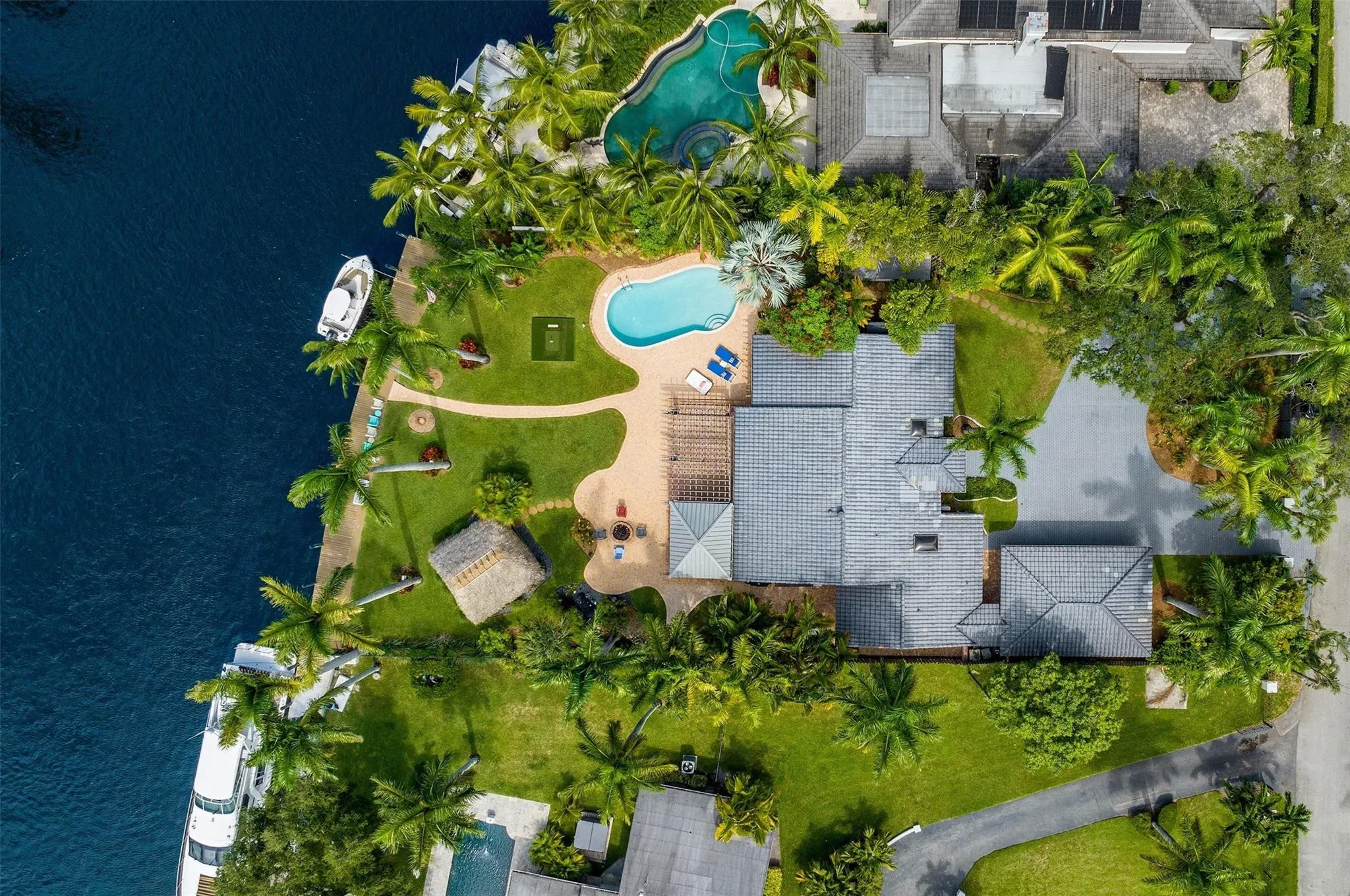 Aerial perspective showcasing extensive waterfront property with direct water access, two distinct swimming pools, a thatched tiki hut, and mature tropical landscaping