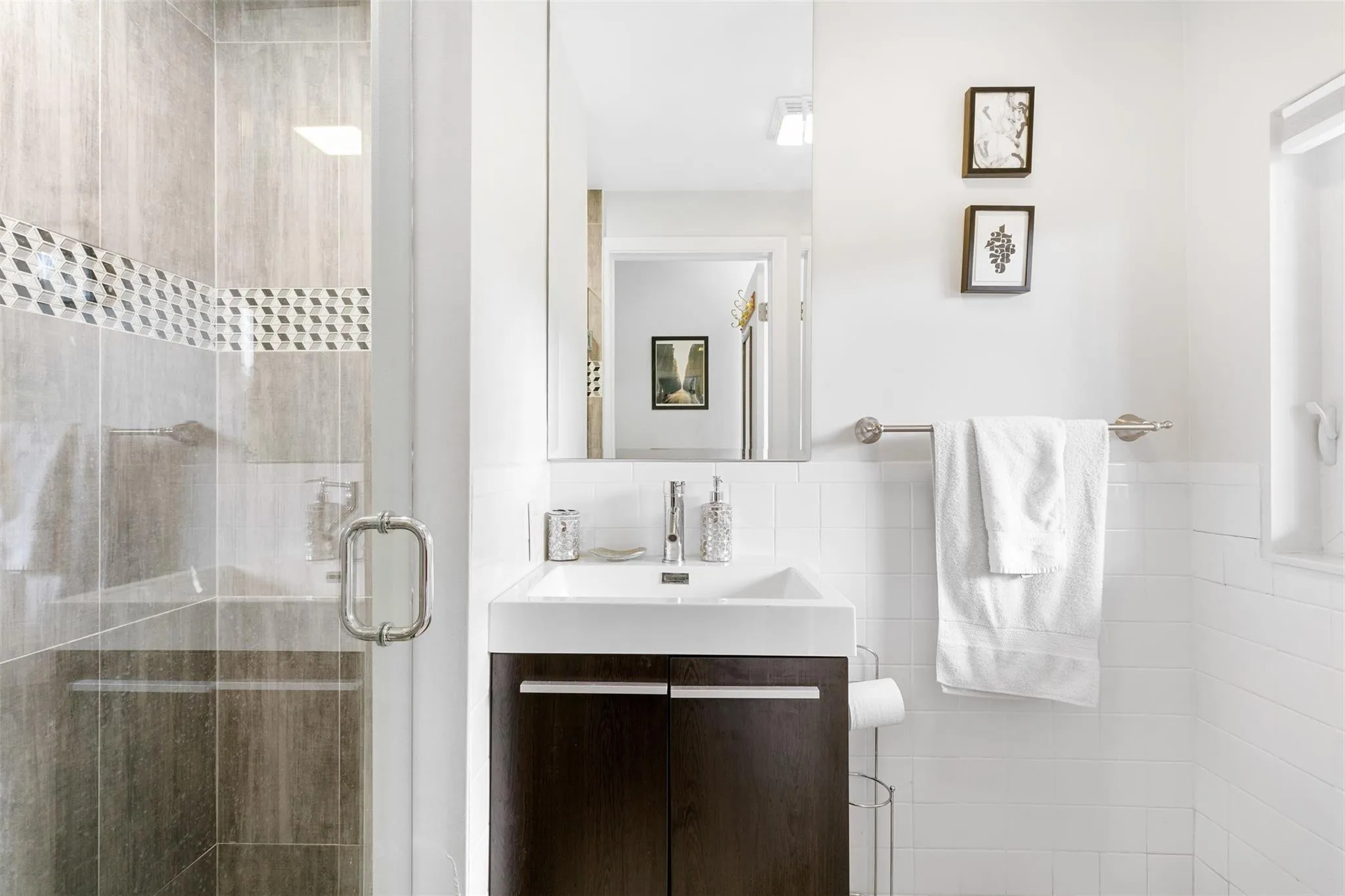 Modern bathroom featuring a dark wood-finish vanity with an integrated white sink, a frameless wall mirror, and a glass-enclosed shower with grey tile and a decorative mosaic accent band