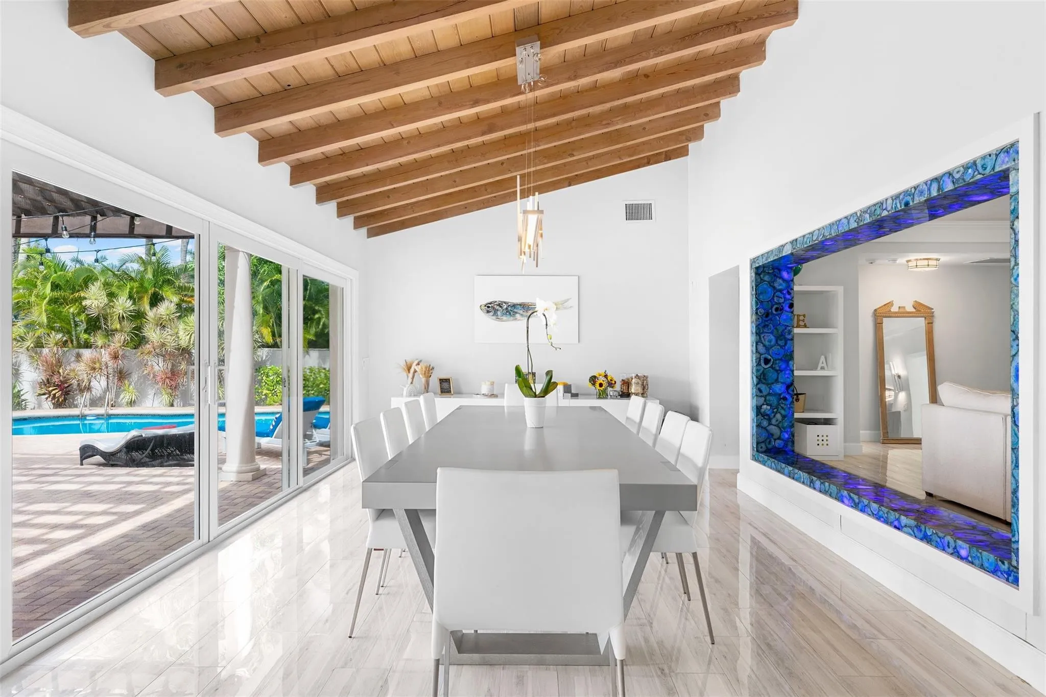 Vaulted wood-beam ceiling with an overhead chandelier, large format tile flooring, and sliding glass doors opening to an outdoor pool area