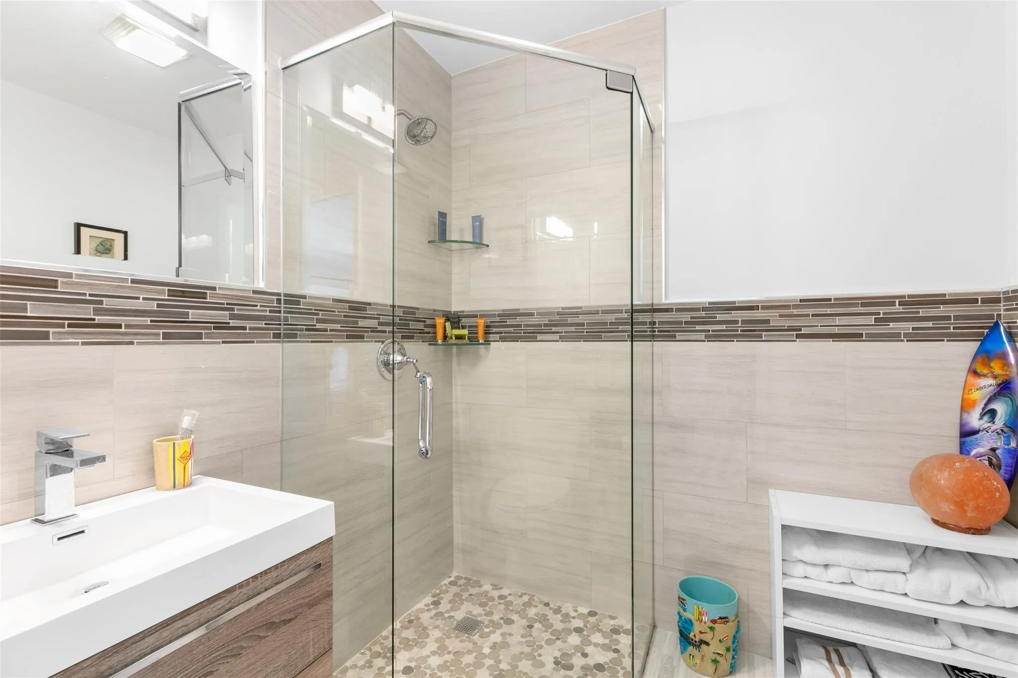 Glass-enclosed shower with pebble-style flooring and light-toned tile surround featuring a decorative mosaic band