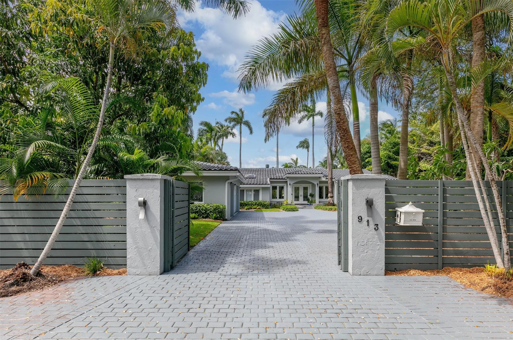 Gated entry featuring a herringbone-patterned paver driveway, concrete pillars with integrated lighting, a horizontally slatted fence, and mature palm trees