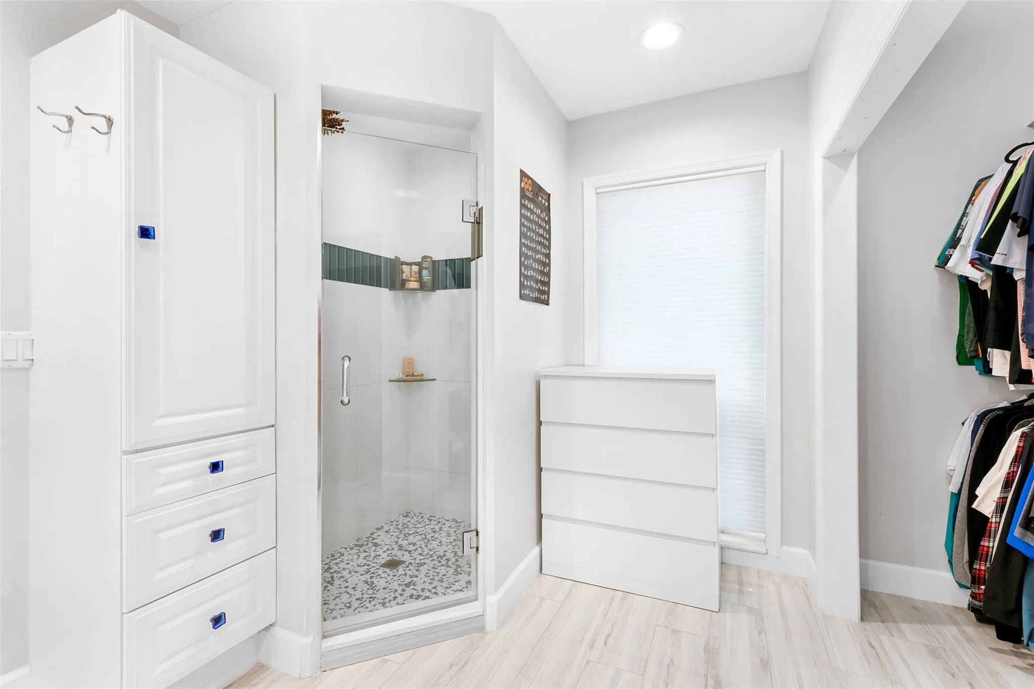 Glass-enclosed shower with pebble-style floor and decorative tile band, alongside a tall white storage cabinet with drawers and an additional white dresser