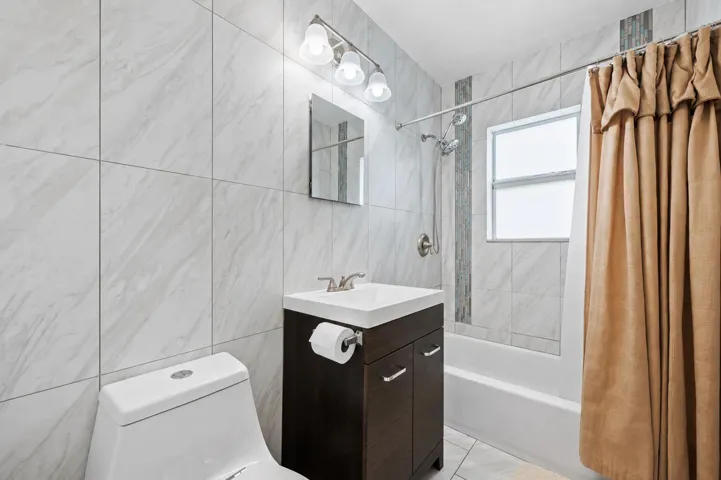 Bathroom featuring white ceramic wall tiling, a dark wood-finish vanity with a white countertop sink, a mirrored medicine cabinet, a three-bulb wall sconce, and a standard white toilet