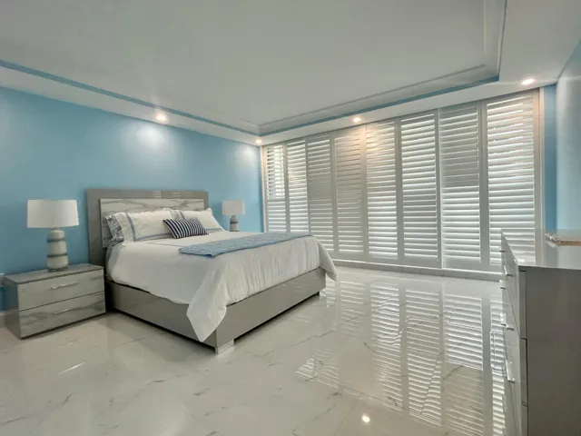 Bright, light-filled bedroom featuring soft blue accent walls, polished light-toned stone flooring, and floor-to-ceiling plantation shutters with integrated lighting