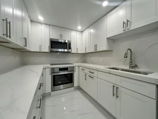 Sleek contemporary kitchen featuring white shaker-style cabinetry with brushed metal hardware, white quartz countertops, stainless steel built-in microwave and wall oven, an undermount sink with modern gooseneck faucet, and marble-look tile backsplash