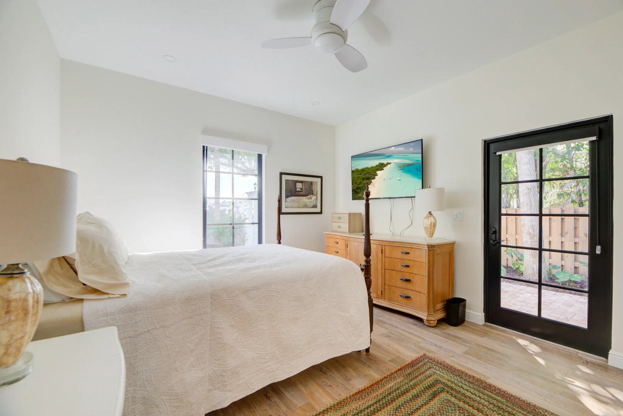Bedroom featuring wood-finish flooring, a ceiling fan, a window with dark framing, and a glass-paned door opening to an outdoor patio with a privacy fence
