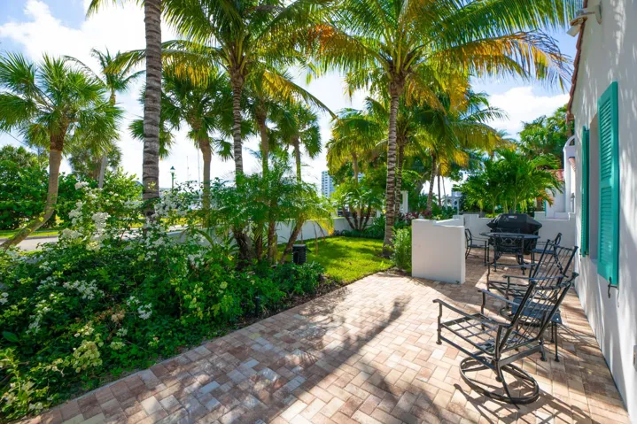 Brick paver patio and walkway featuring a white stucco exterior with teal louvered shutters, mature palm trees, and established landscaping