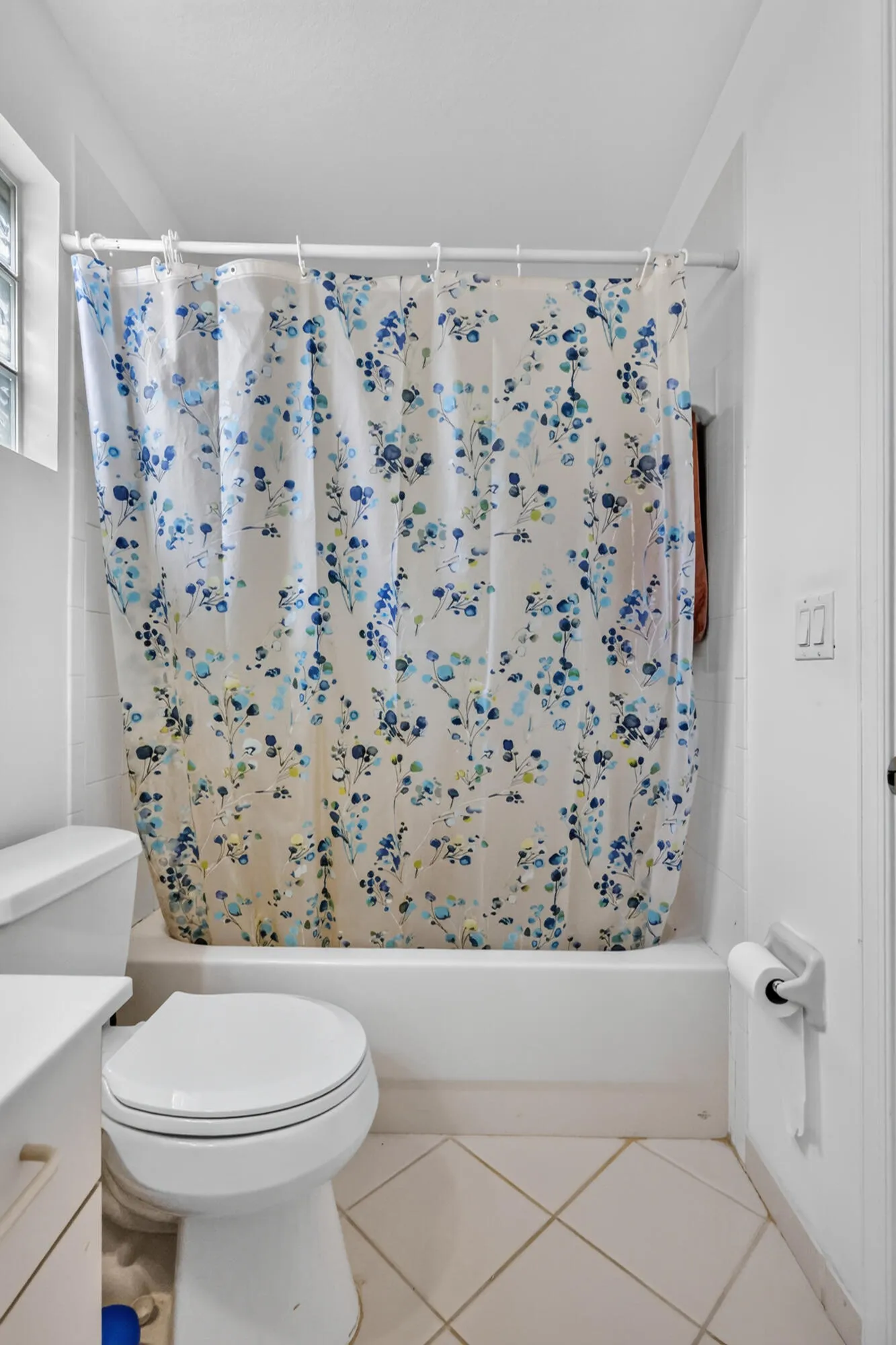 White-tiled bathroom featuring a built-in tub with shower, glass block window, and light-toned floor tiles