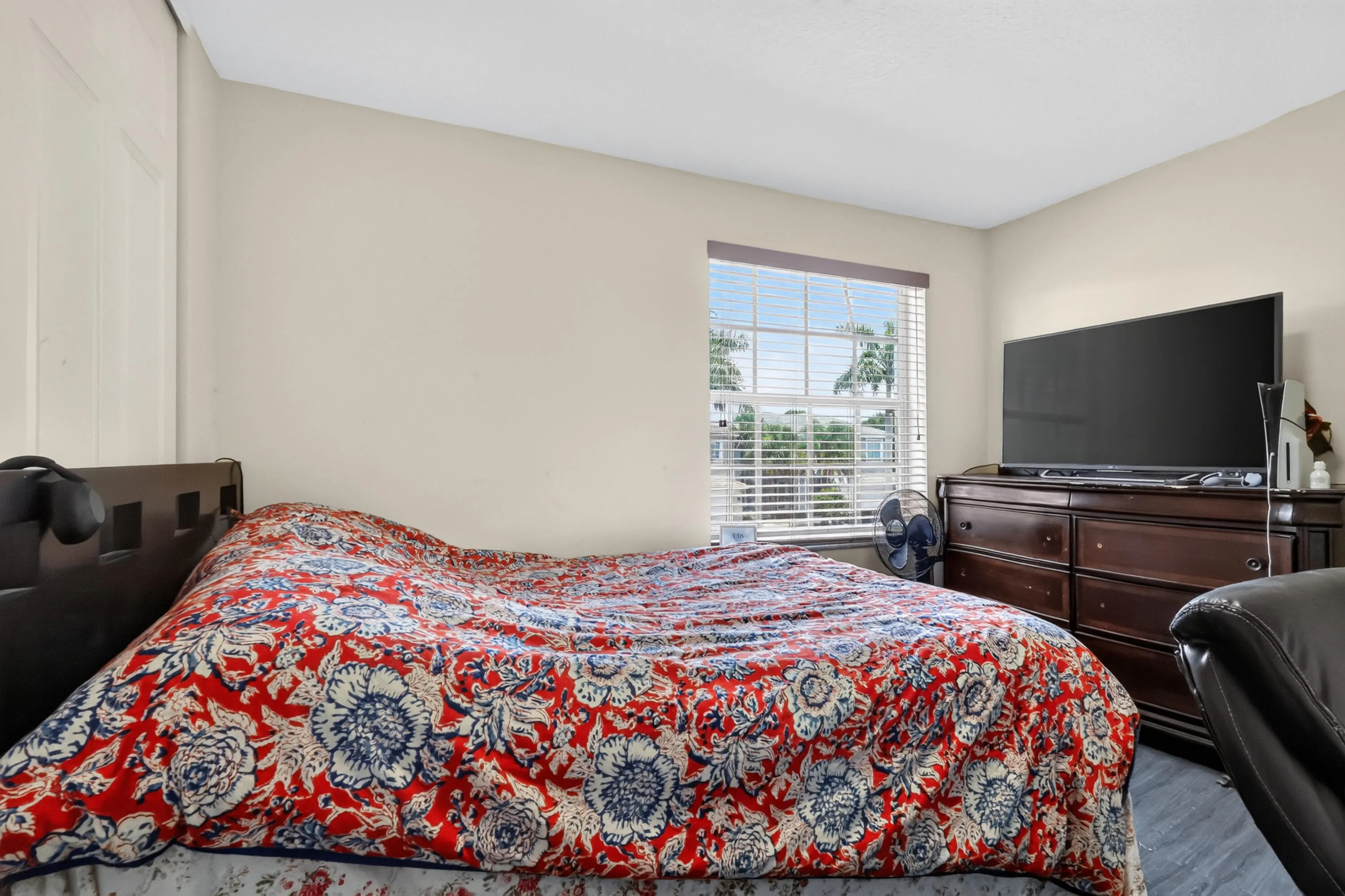 Comfortable bedroom featuring a large bed with decorative bedding, wood-finish dresser with integrated television mounting, dark upholstered seating, and abundant natural light from a multi-pane window with plantation shutters