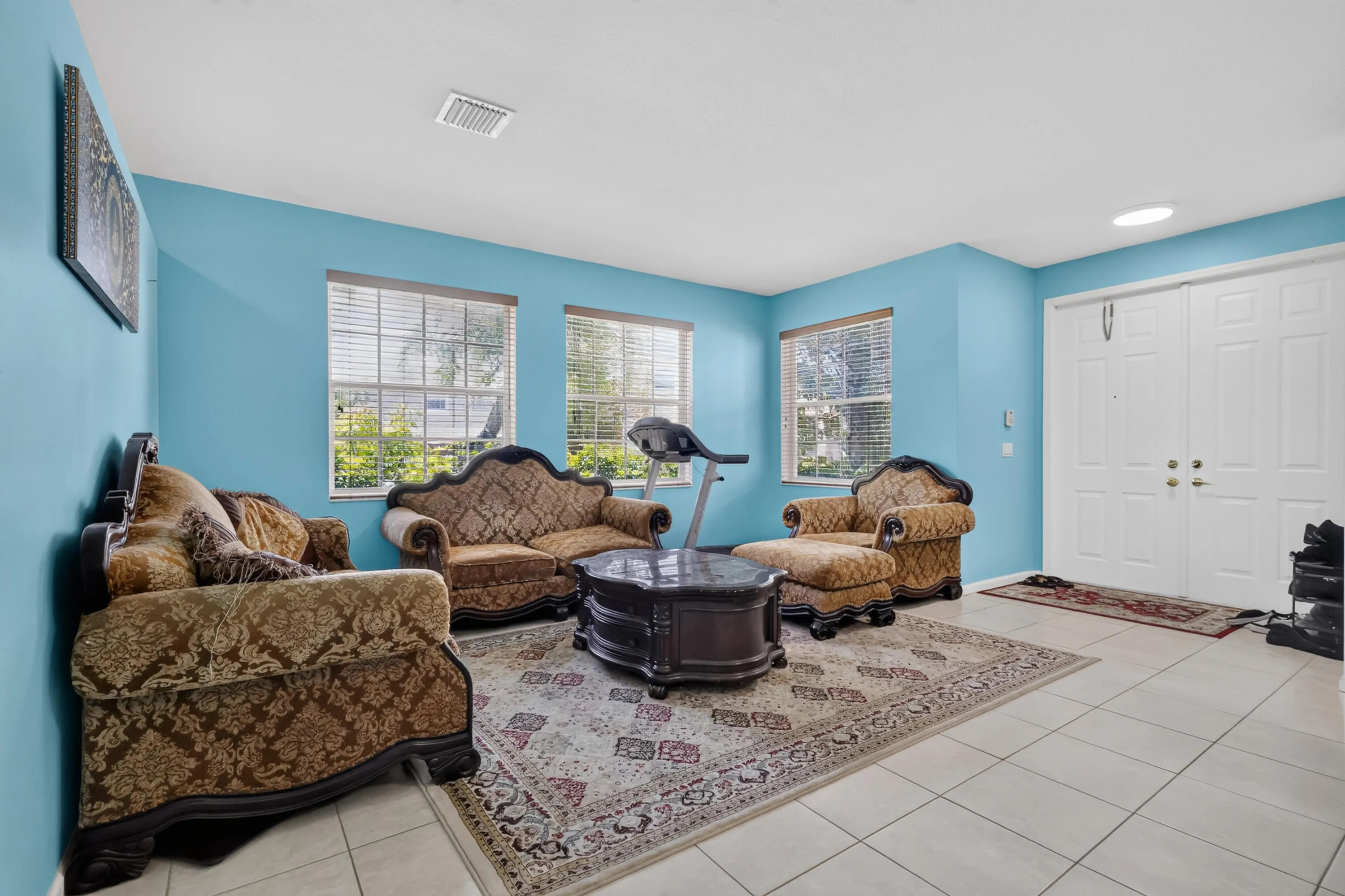 Spacious living area featuring light-toned tile flooring, three large windows with horizontal blinds, and a white double entry door