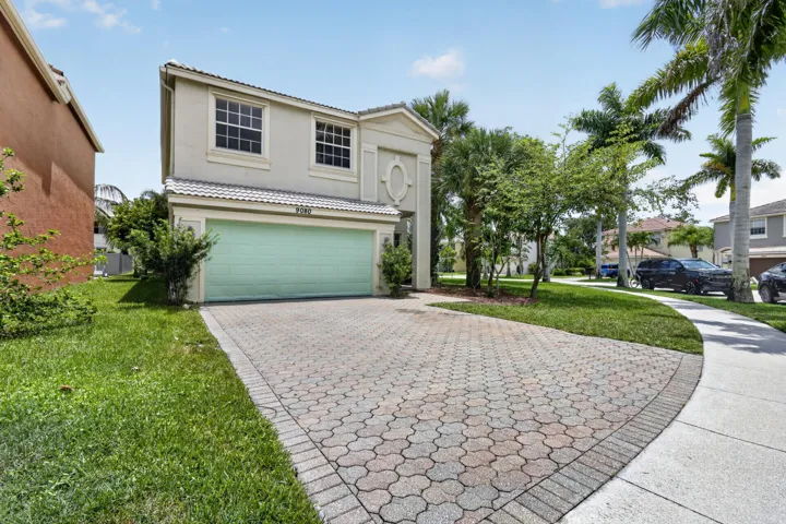 Two-story residence with a light-colored exterior, featuring a two-car garage with mint-green doors, paver driveway with geometric pattern, manicured lawn, and mature landscaping including palm trees and tropical vegetation