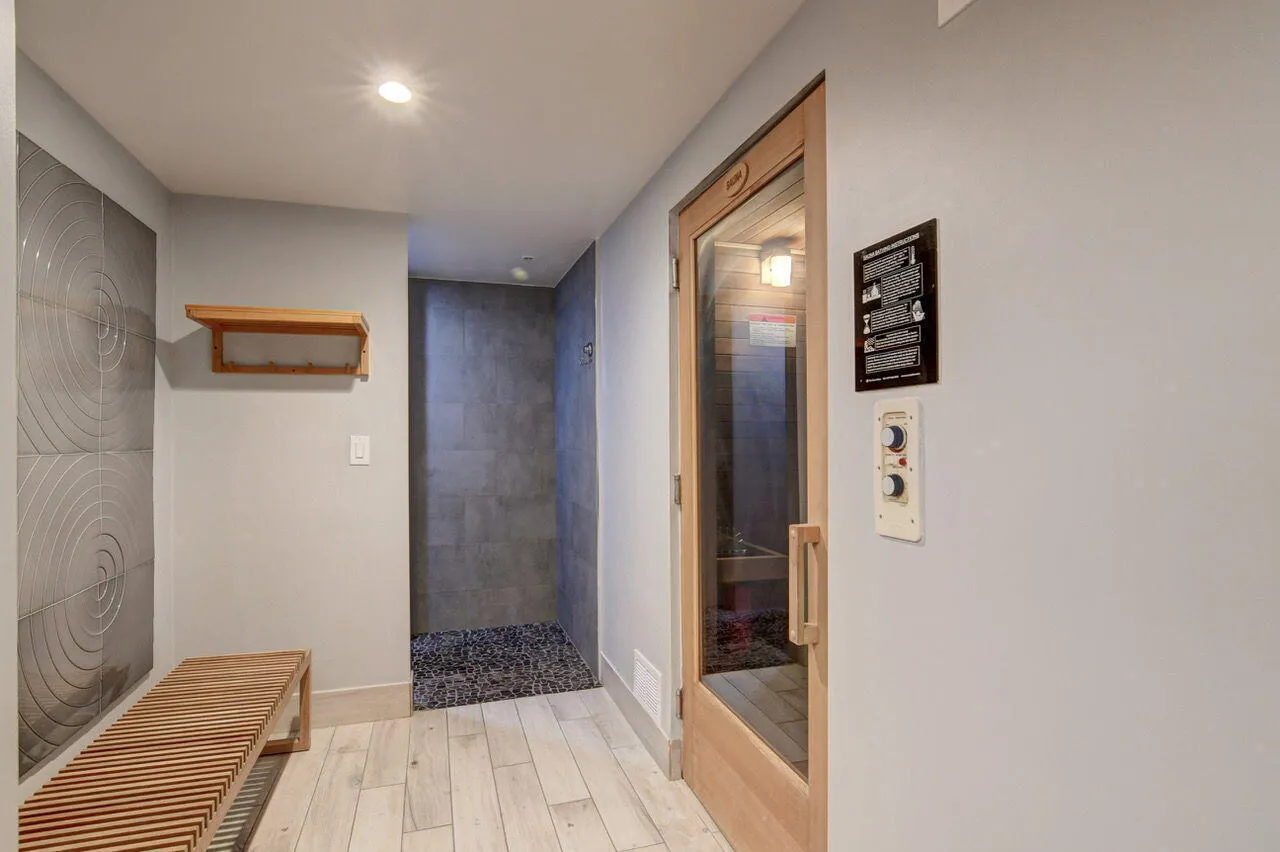 Dedicated sauna room with a clear door and exterior controls, featuring wood-finish flooring, a spa-style shower with dark pebble-stone base and wall tiling, a slatted wood bench, and textured wall panels