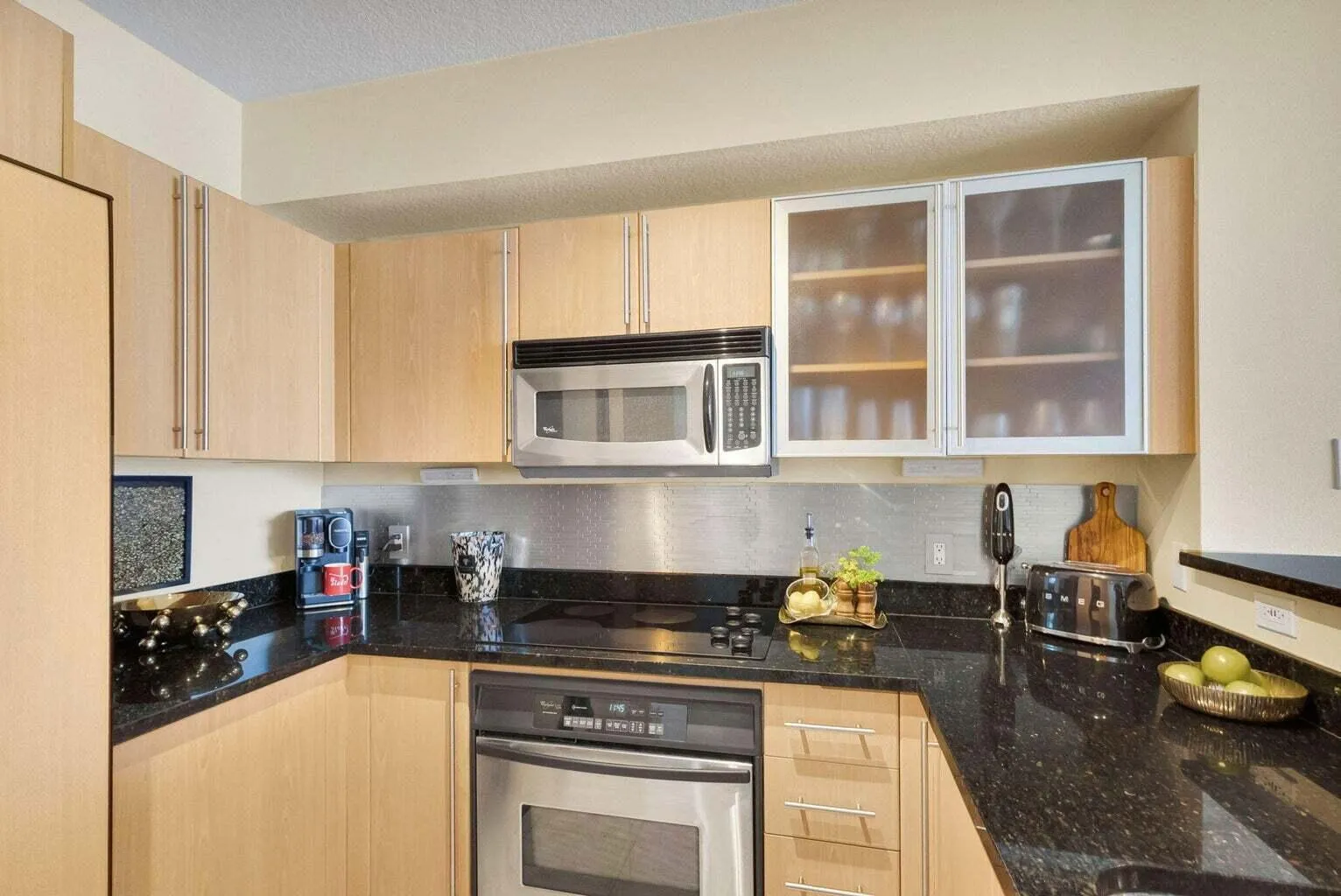 Contemporary kitchen featuring wood-finish cabinetry with brushed nickel pulls, black speckled countertops, stainless steel appliances, and a coordinating metal backsplash