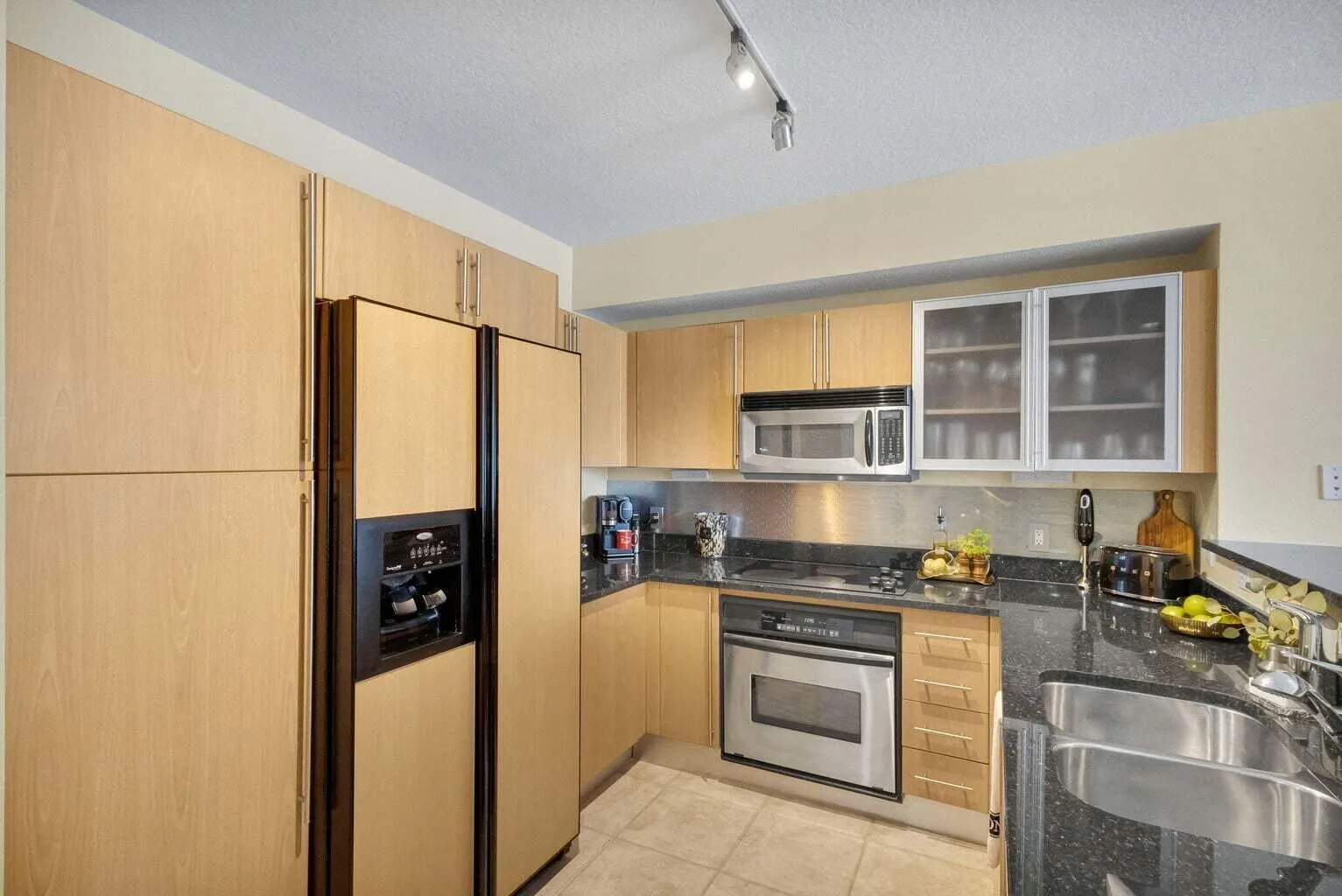 Kitchen featuring light wood-finish cabinetry, dark stone countertops, stainless steel appliances, a stainless steel backsplash, and dual stainless steel sinks