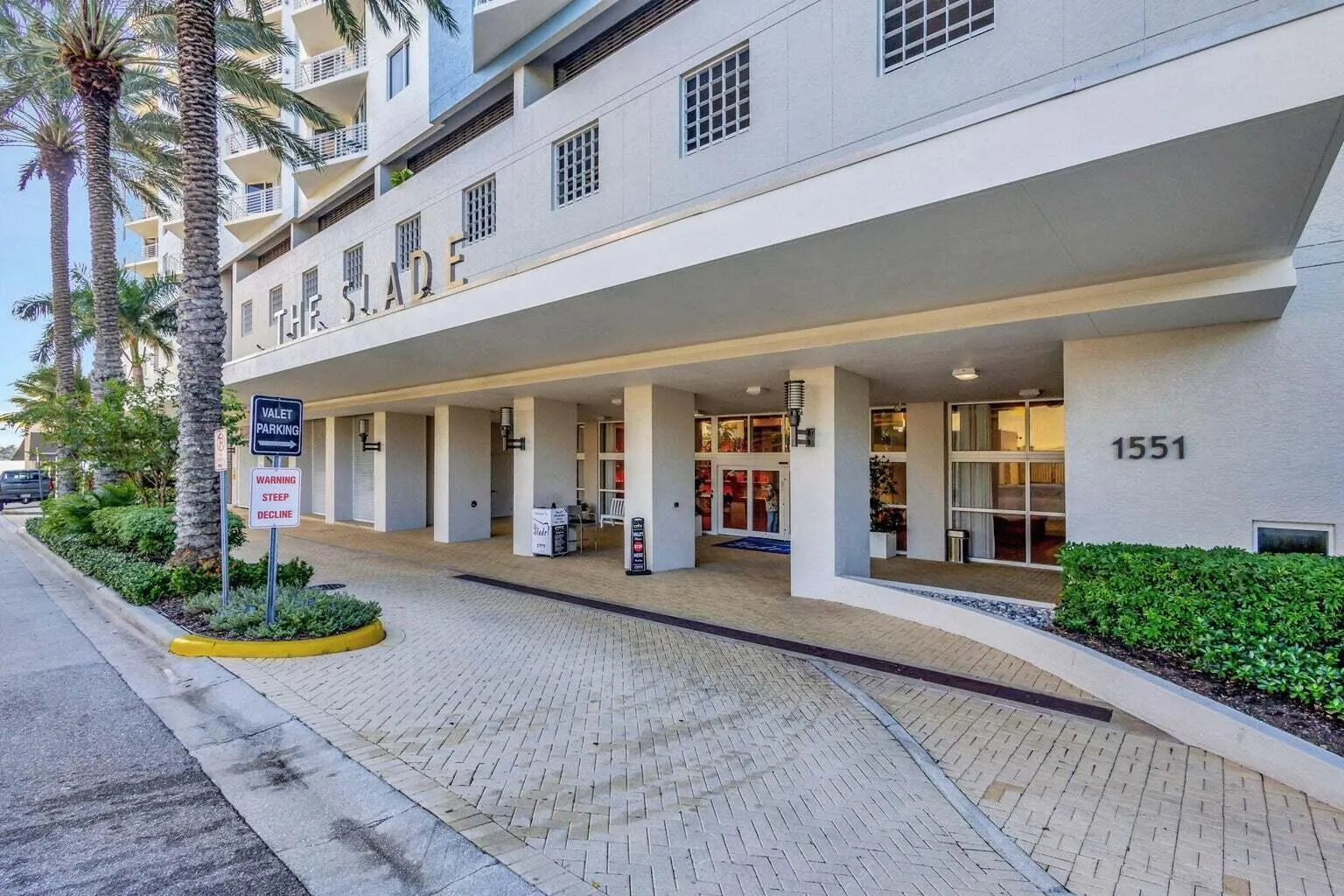 Modern building entrance featuring a patterned paver driveway, lush landscaping, prominent building signage, and a covered drop-off area