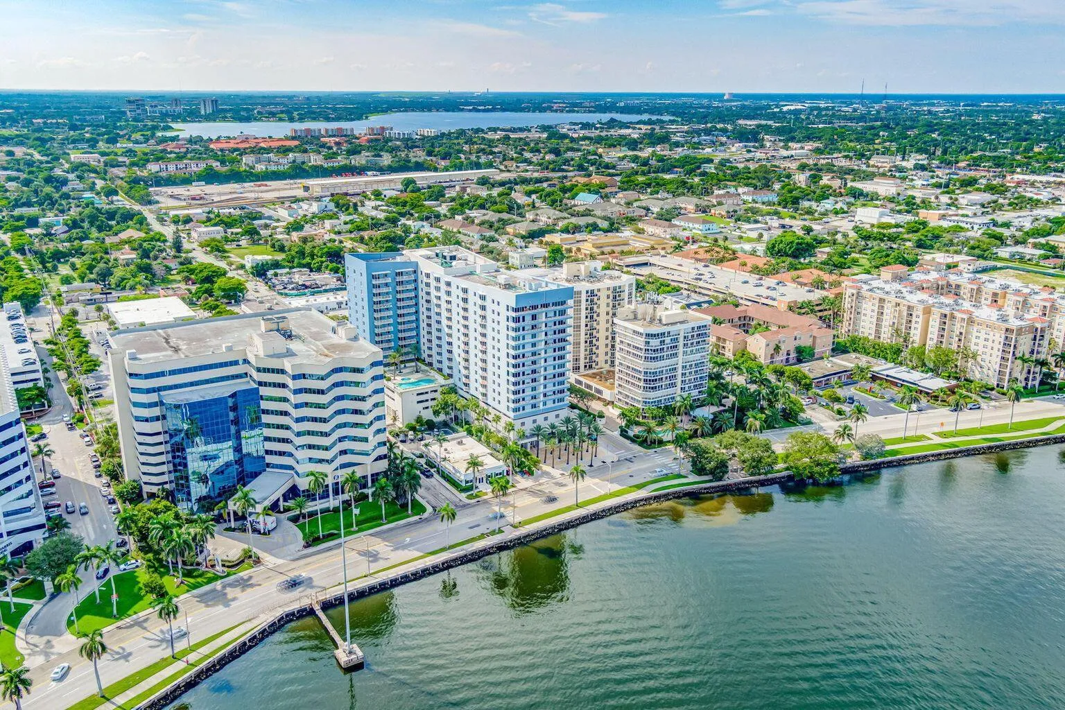 Waterfront cityscape featuring multiple high-rise buildings, extensive green landscaping, a prominent waterway, and a distant body of water