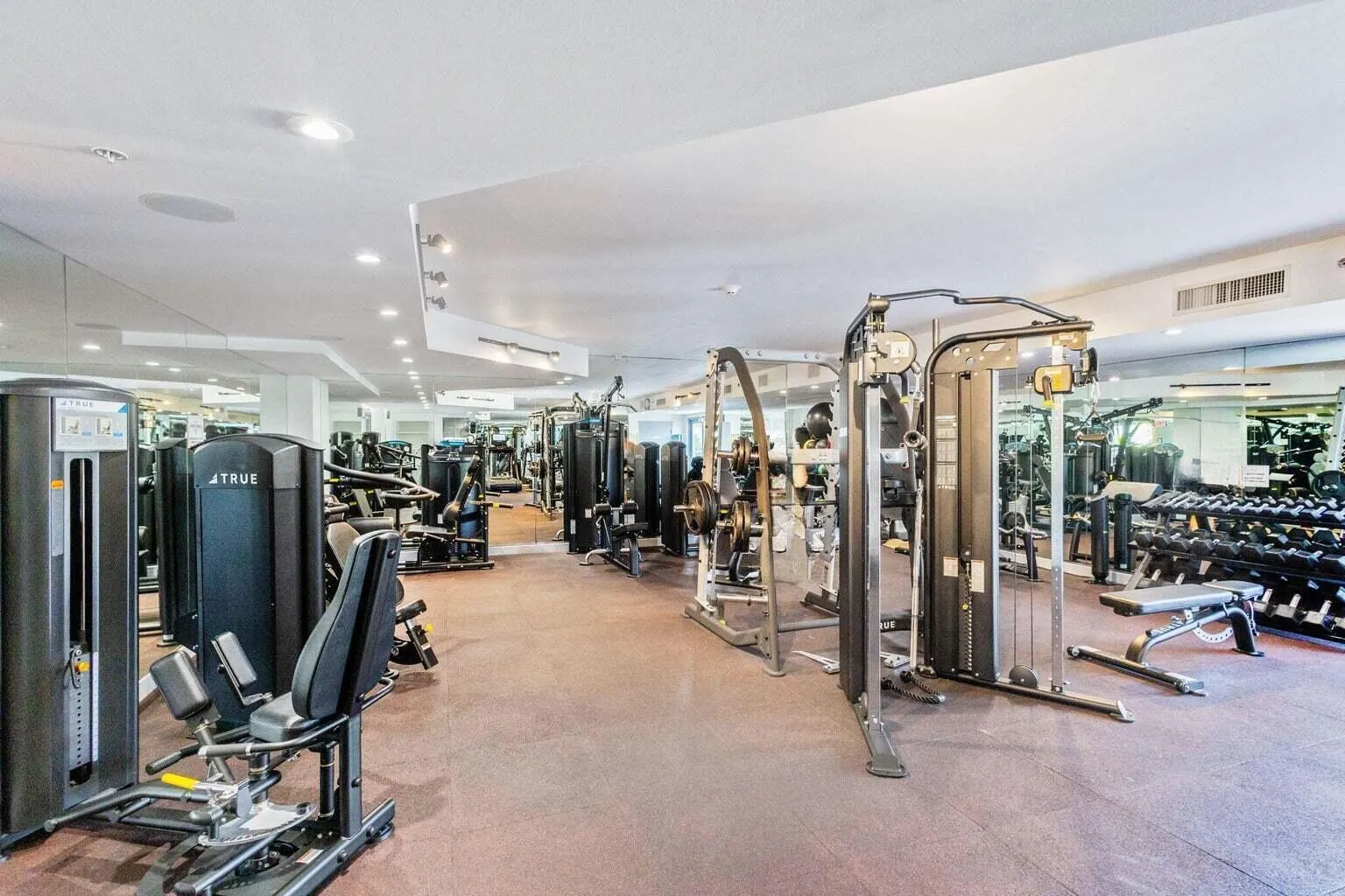 Equipped fitness center featuring various strength training machines, free weights, mirrored walls, recessed lighting, and a durable floor surface