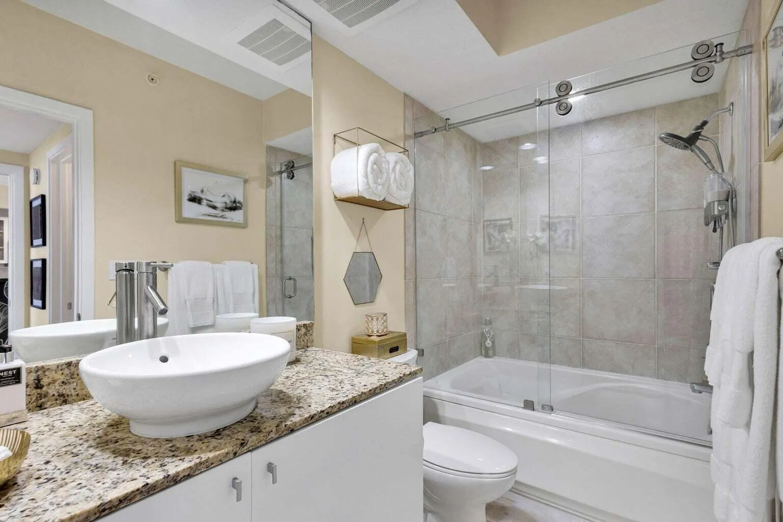 Bathroom featuring a white vessel sink set on a speckled countertop, a mirrored vanity, a commode, and a tub with a sliding glass shower door and tiled surround