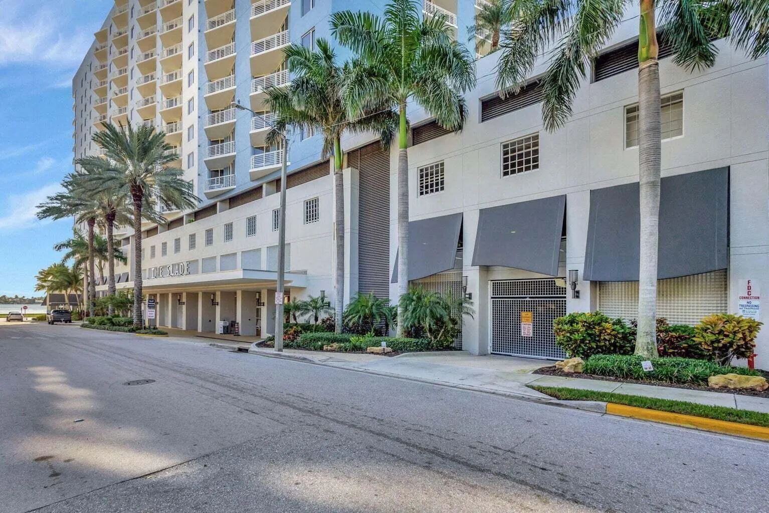 Waterfront high-rise building featuring multiple levels of balconies, ground-floor retail spaces, a gated parking entrance with awning coverage, and tropical landscaping with mature palm trees