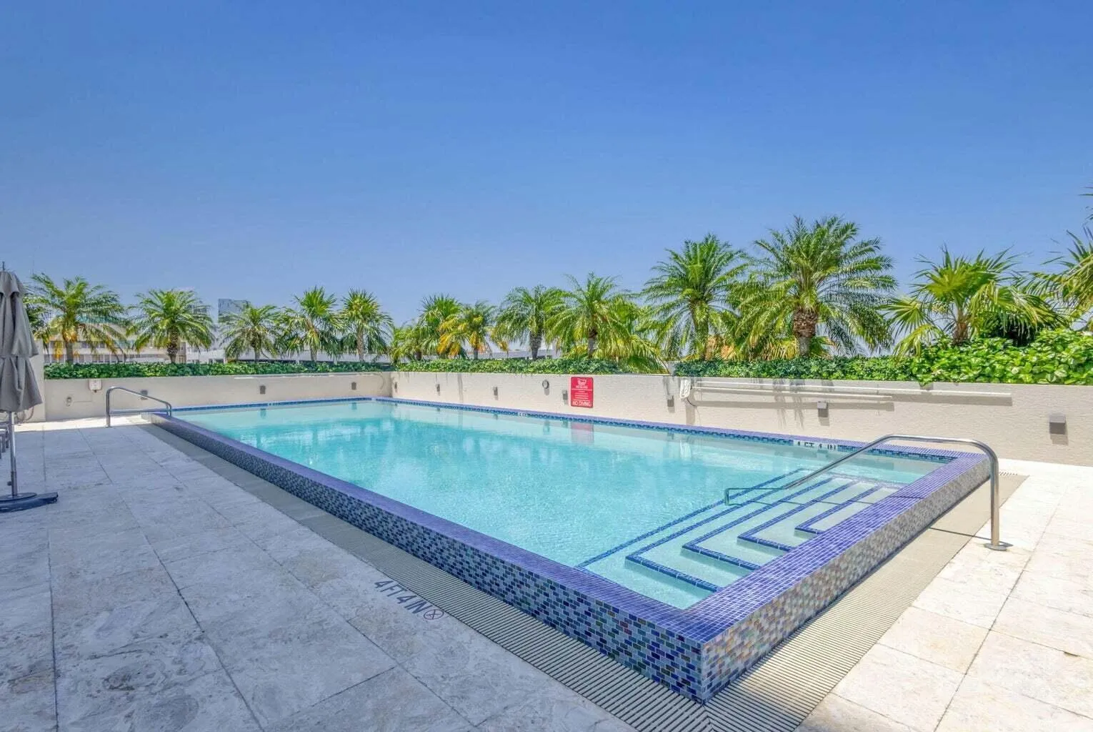 Rectangular swimming pool featuring a blue mosaic tile border and integrated steps, surrounded by light-colored tile decking and privacy wall