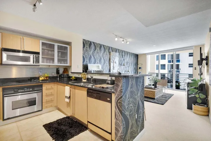 Open concept living area featuring a kitchen with light wood cabinetry, stainless steel appliances, and dark countertops