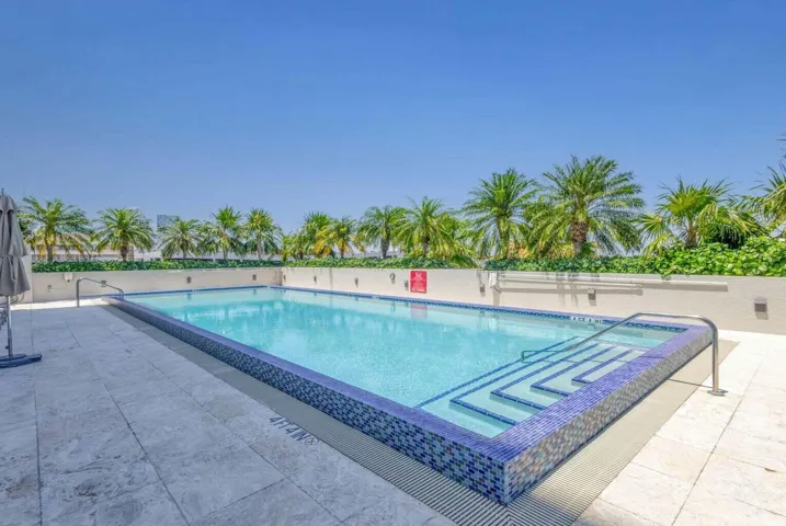 Rectangular swimming pool featuring a blue mosaic tile border and integrated steps, surrounded by light-colored tile decking and privacy wall