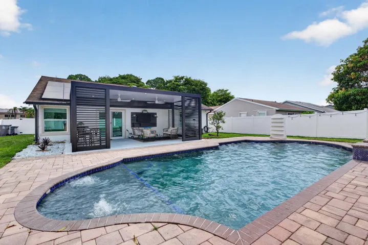 Travertine-style paver patio surrounding a freeform pool, featuring a covered outdoor living area with ceiling fans and an integrated media wall