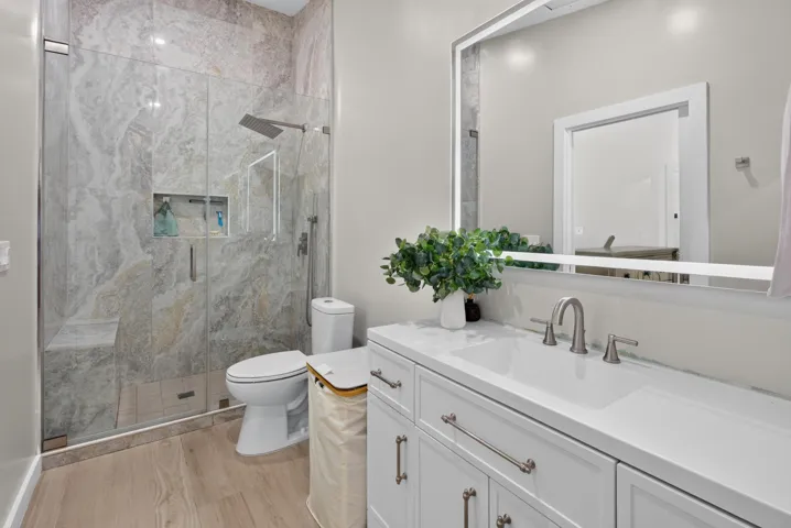 Spa-inspired primary bathroom featuring a frameless glass-enclosed walk-in shower with marble-look tile surround, a white vanity with dual sink configuration and polished quartz countertop, chrome fixtures, and wood-finish flooring throughout