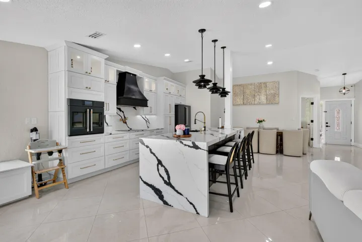 Sophisticated open-concept kitchen and dining area featuring a striking waterfall-edge island with marble-look countertops, white cabinetry with integrated appliances including a black range hood, polished light-stone flooring, pendant lighting, and black metal dining chairs