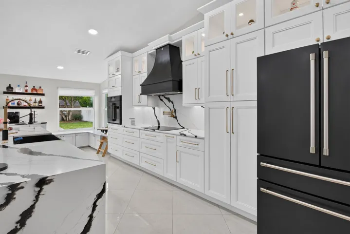 Gourmet kitchen featuring a large central island with waterfall-edge countertop, white shaker cabinetry, integrated black appliances, a black range hood, and light tile flooring