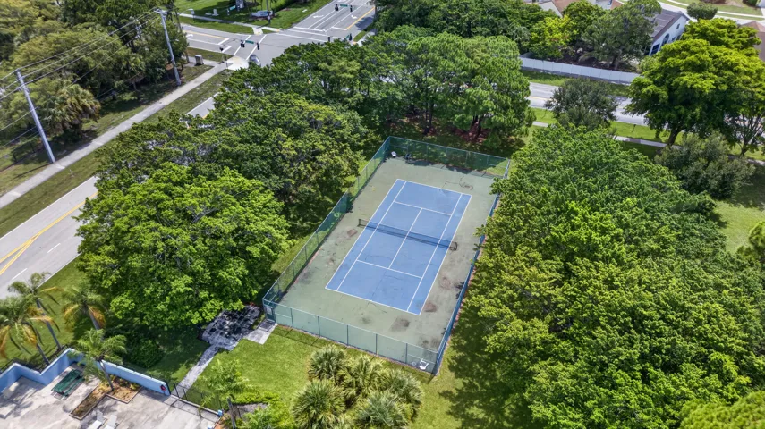 Fenced tennis court featuring a blue playing surface and green surrounds, bordered by mature trees and lush landscaping