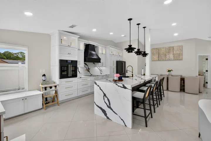 Gourmet kitchen featuring a waterfall island with integrated seating, white cabinetry, integrated double ovens, a black range hood, and light-toned tile flooring